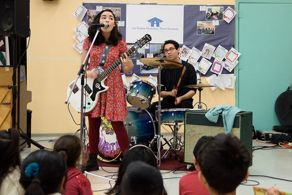 Una joven en un vestido roja toca la guitarra y canta en un micrófono. Los niños están sentados en el suelo.