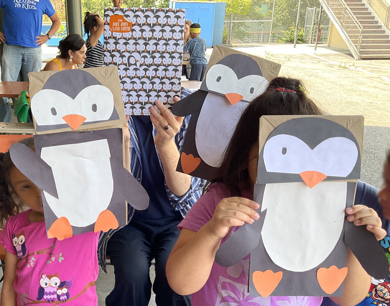 Un grupo de niños levantan sus manualidades de pájaro de papel, mientras una lectora voluntaria levanta un libro con tema pájaro.