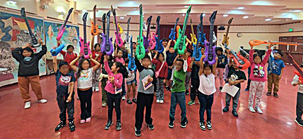 Kids holding up their new inflatable guitars after enjoying music by JEONG at Charles White Elementary
