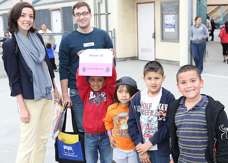Dos voluntarios y cuatro niños están de pie afuera de la escuela, todos sonriendo. Un niño sostiene un cartel que dice 'Room 12, Kindergarten,' mientras un voluntario sostiene una bolsa con útiles.